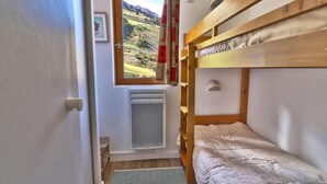 Een kleine, smalle kamer met een stapelbed, een radiator, een raam met uitzicht op een bergachtig landschap en een deur.