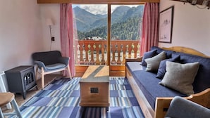 Een gezellige woonkamer met een bank, een stoel, een kleine tafel en een verwarming. Door het raam is een balkon met houten reling en uitzicht op bergen zichtbaar.