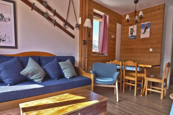 A cozy room with a wooden wall, a blue sofa with cushions, a wooden dining table with chairs, and a window with a pink curtain.