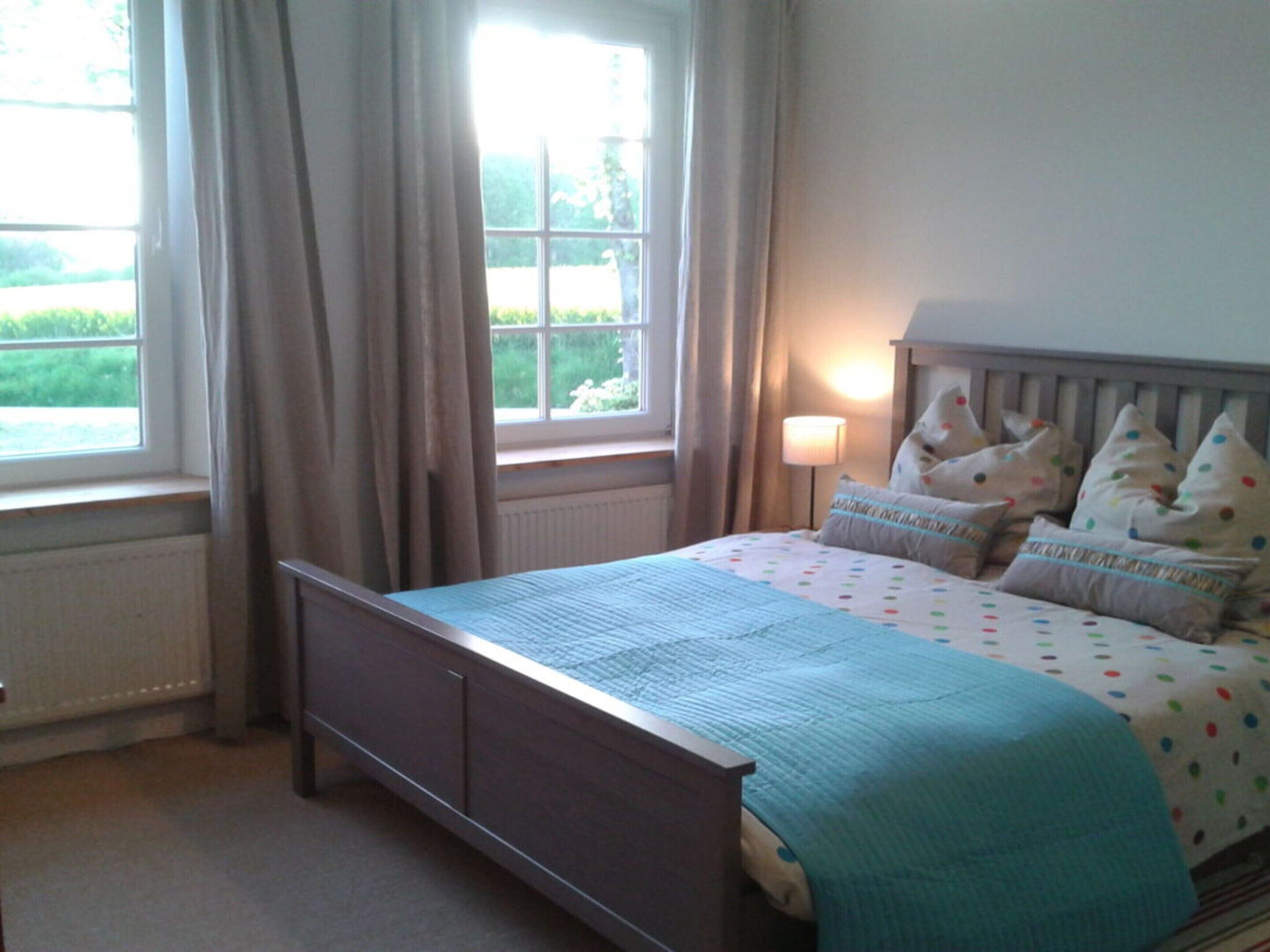 A bedroom with a bed, a window with curtains, a bedside lamp, and a view of greenery outside.