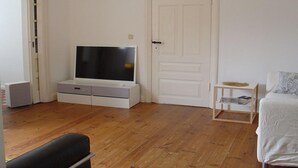 Une pièce avec un plancher en bois, une porte blanche, un téléviseur posé sur un meuble blanc, une petite table d’appoint en bois avec un magazine, et un lit recouvert d’une couverture blanche.