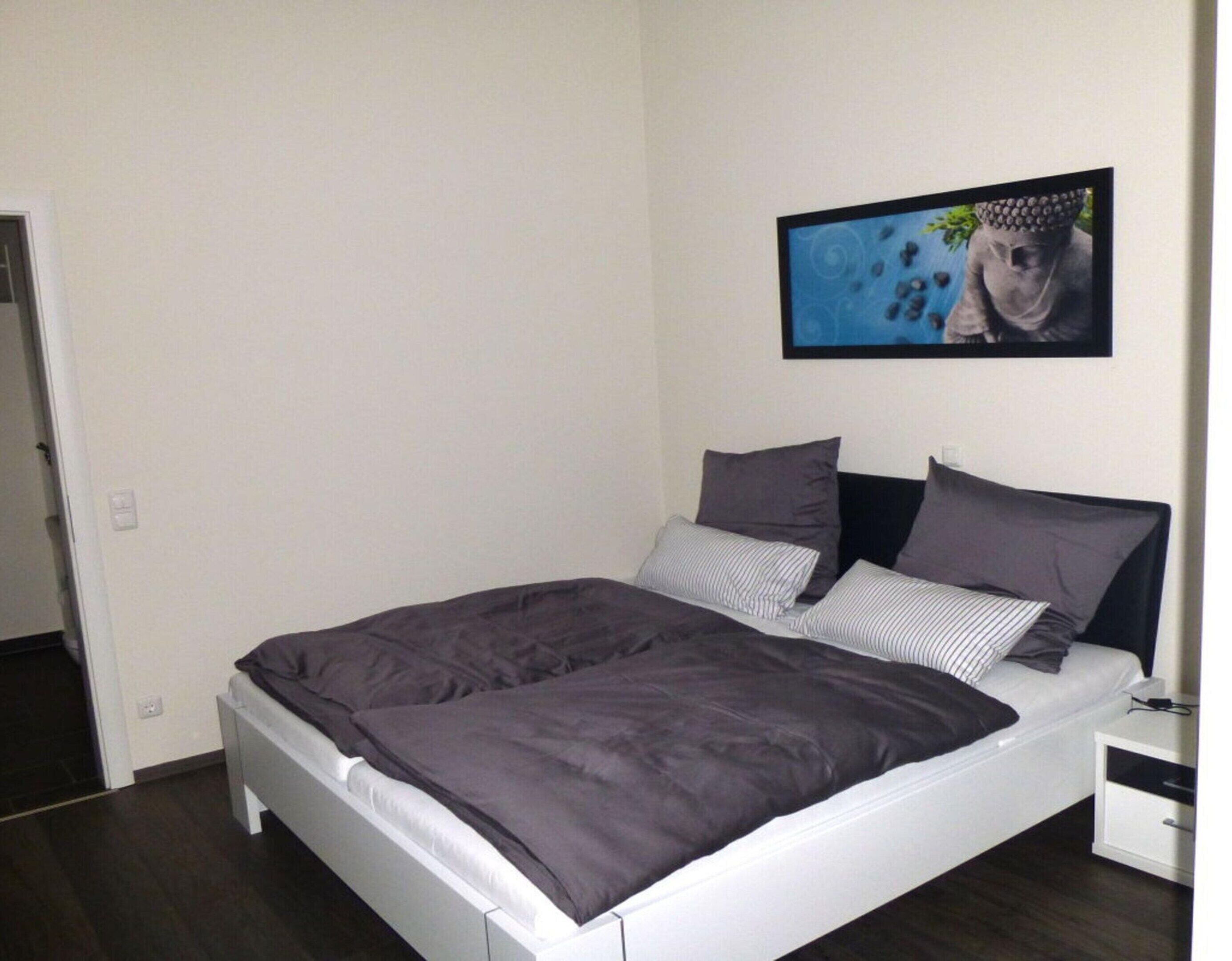 Ein ordentlich bezogenes Bett mit einer dunkelgrauen Tagesdecke und Kissen, einem weißen Kopfseitenverkleidungen und einem kleinen Nachttisch mit einer Lampe.