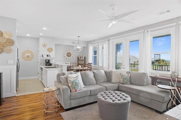 A modern living room with a grey sectional sofa, a round ottoman, and a dining area with a table and chairs.