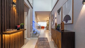 A modern hallway with a wooden reception desk labeled "DREAM," a staircase, and a dining area in the background.