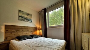 Ein Schlafzimmer mit Holz-Kopfteil, einem großen Bett mit weißer Bettwäsche, einem Nachttisch mit Lampe, einer Kommode und einem Fenster mit Vorhängen.