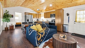A spacious living room with a dark blue sofa, wooden ceiling, and a kitchen area with wooden cabinets and appliances.