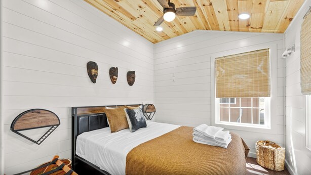A bedroom with a wooden ceiling, a bed with a headboard, a window with blinds, and wall-mounted artwork.
