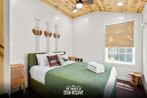 A bedroom with a green bedspread, a wooden ceiling, and a window with blinds.