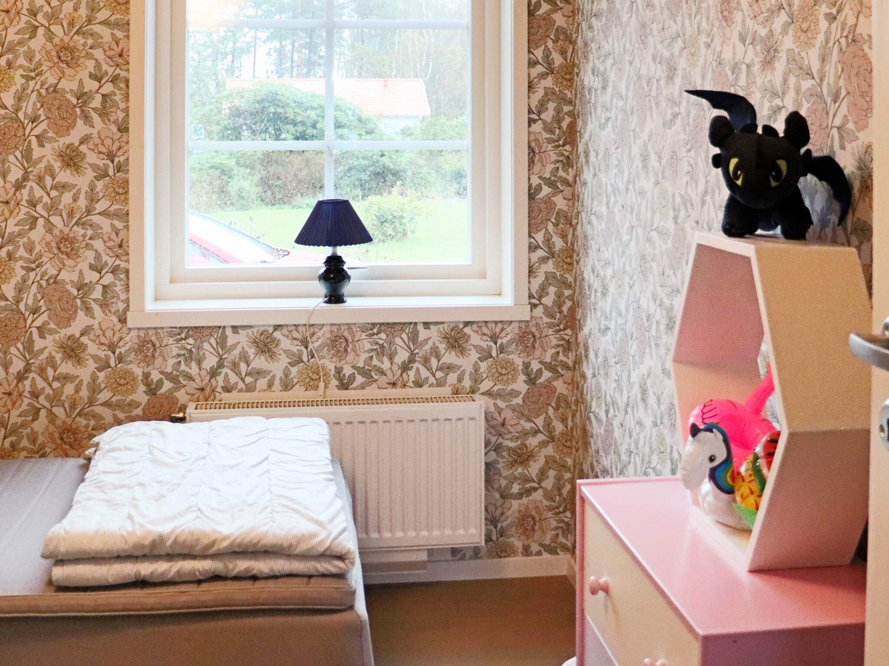 Ein Schlafzimmer mit Fenster, Heizkörper, einem Bett mit weißer Decke und einer Kommode mit Stofftier.