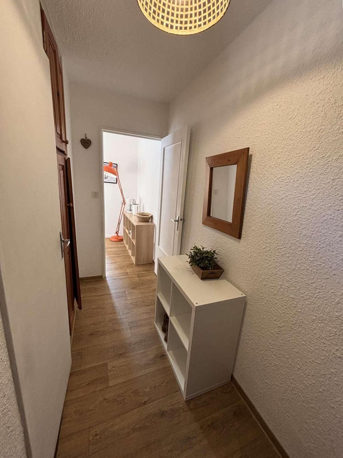 Eine Diele mit Holzboden, einem weißen Konsolentisch mit Pflanze, einem Spiegel und einer Tür zu einem Zimmer mit Bett und Fenster.