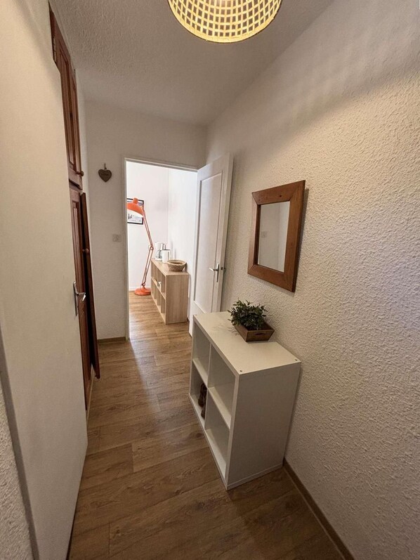 A hallway with a wooden floor, a white console table with a plant, a mirror, and a door leading to a room with a bed and a window.