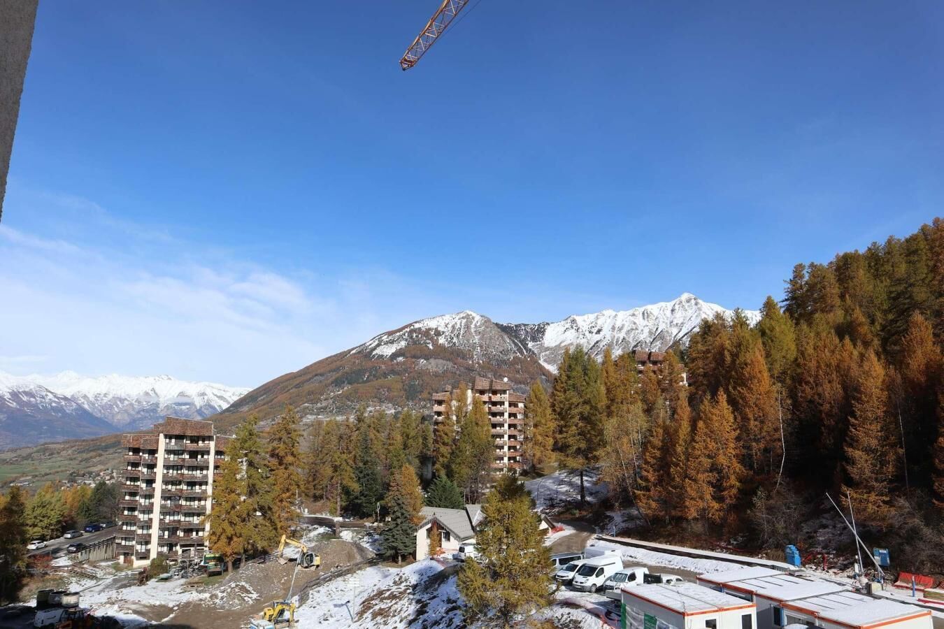 A mountainous landscape with snow-capped peaks, a forested area, and several buildings including a multi-story hotel with balconies.