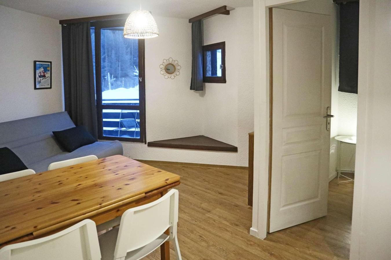 A cozy room with a wooden dining table, white chairs, a sofa, a small window with a view of trees, and a door leading to another room.