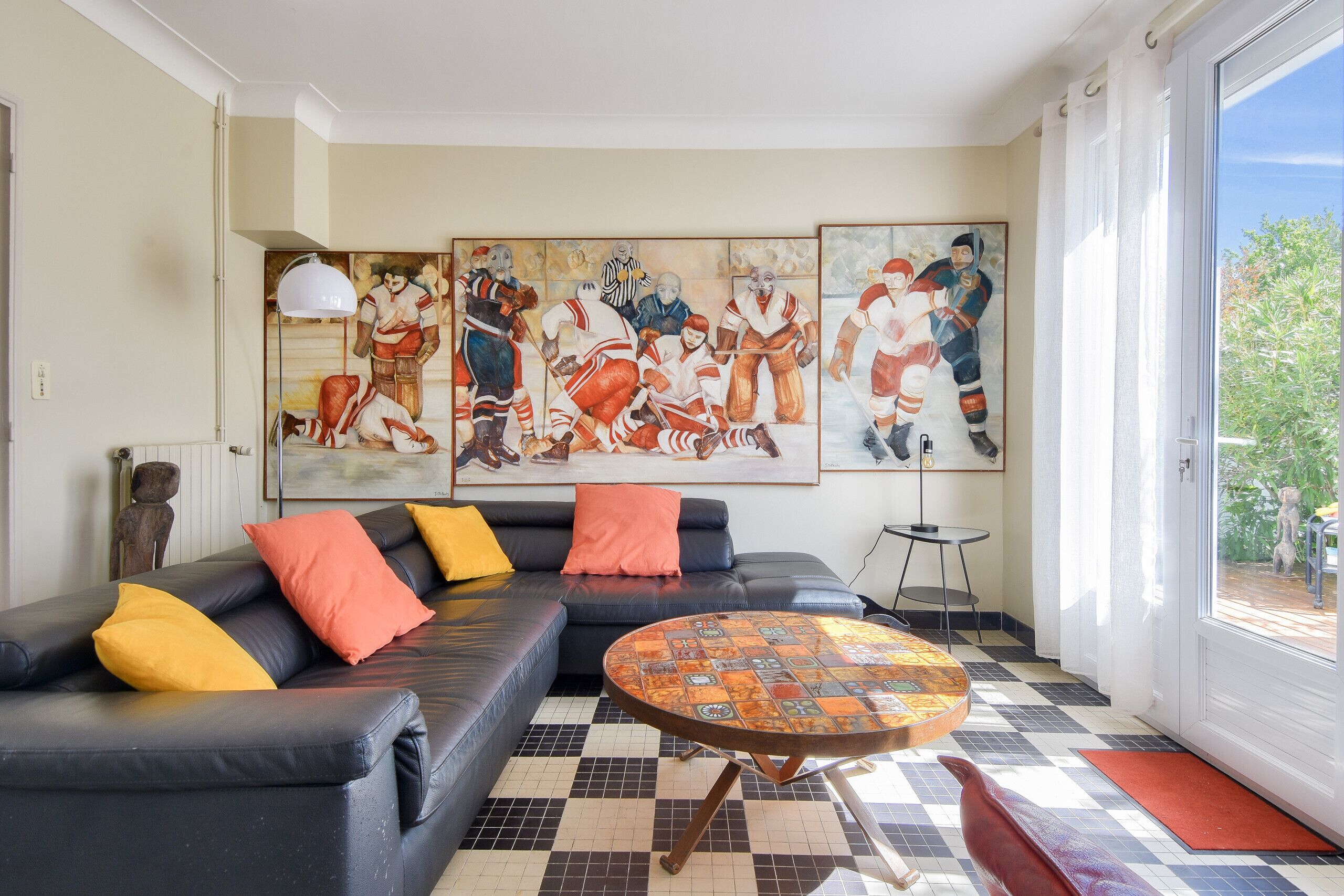 A living room with a black leather sofa, colorful cushions, a patterned coffee table, and large sports-themed paintings on the wall.