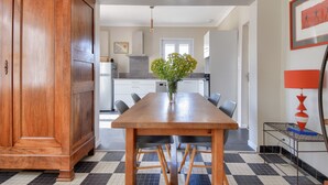 A dining area with a wooden table, chairs, a vase with flowers, and a kitchen in the background.
