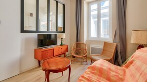 A hotel room with a large mirror, a television on a wooden cabinet, a wicker chair, a bed with a plaid blanket, and a window with curtains. - Getaway 150m from the beach in Les Sables d'Olonne (Les Sables-d´Olonne)