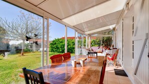 Une terrasse couverte avec une table et des chaises en bois, de grandes fenêtres et une vue sur le jardin et les bâtiments voisins.