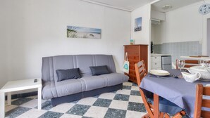 Ein Zimmer mit grauem Sofa, einem kleinen weißen Tisch, einem Schachbrettboden, einem Holzkabinett und einem Essbereich mit Tisch für zwei Personen.