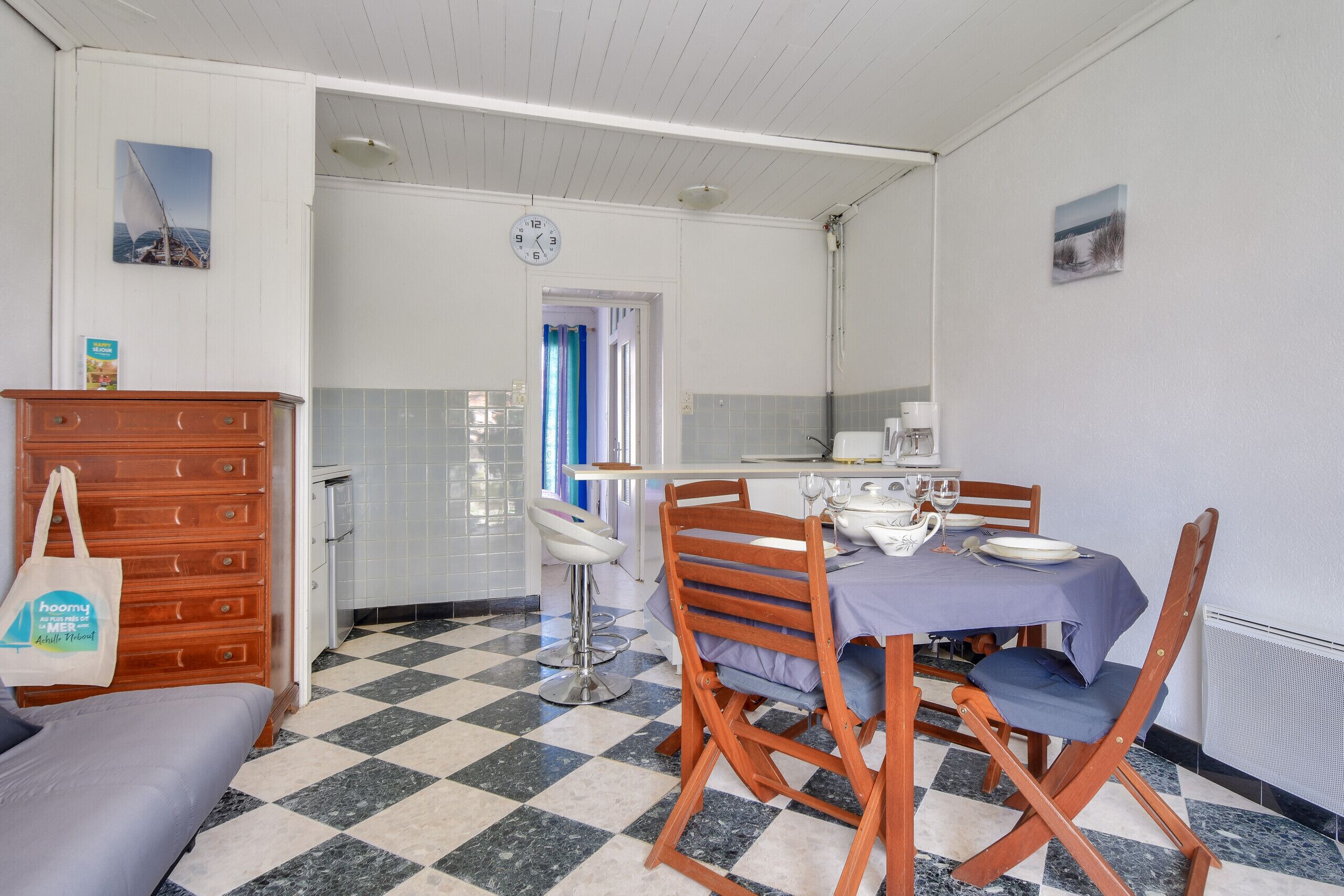 Un comedor con piso a cuadros, sillas de madera, una mesa con mantel azul y un aparador de madera con un bolso encima.