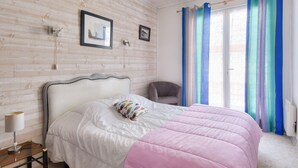 Ein Schlafzimmer mit einer Holzwand, einem Bett mit einer weiß-rosa Steudecke, einem Sessel und einem Fenster mit blauen Vorhängen.