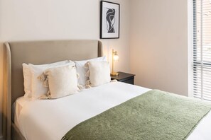 Een net opgemaakt bed met een groene plaid, een bedlamp en een lijst met abstracte kunst aan de muur.