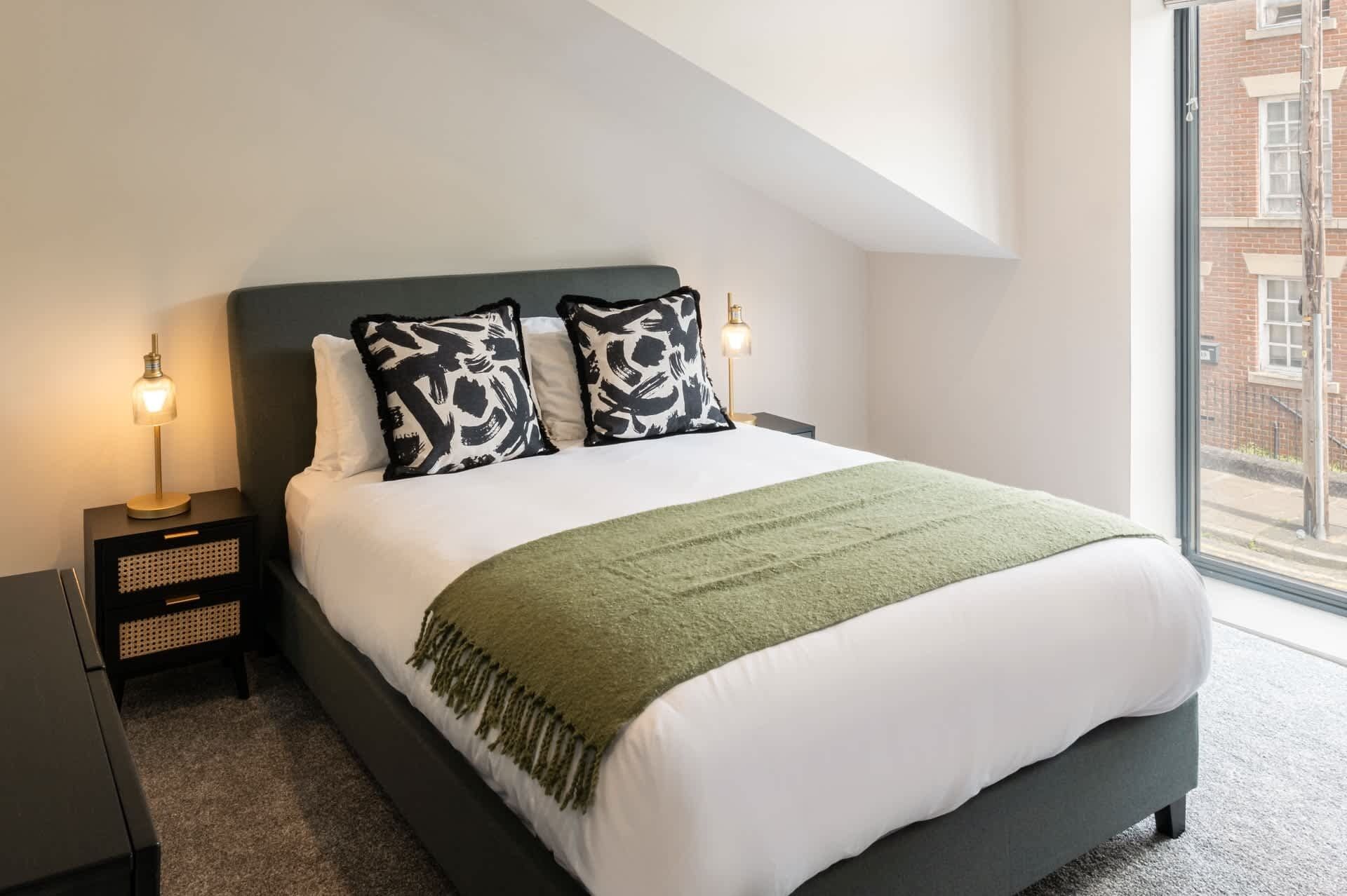 A neatly made bed with a green throw, two bedside lamps, and a view of a brick building through a window.