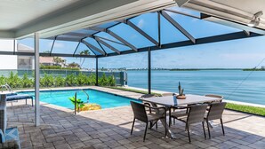 A covered outdoor area with a pool, dining table, and chairs, overlooking a body of water. - Waterfront 3/3 Home with Panoramic Bay Views! (Marco Island)