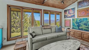 A modern living room with a large window, a grey sofa, and a marble coffee table. - GrandView @ Smoky Ridge: Stunning Mtn Views, Large Deck & Windows | Pet Friendly (Cullowhee)