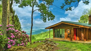 A wooden cabin with a glass door, surrounded by greenery and mountains. - GrandView @ Smoky Ridge: Stunning Mtn Views, Large Deck & Windows | Pet Friendly (Cullowhee)