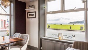 A dining area with a view of a green field and water, a table with chairs, and a hanging light fixture. - Lamlash Bay Hotel (Isle of Arran)