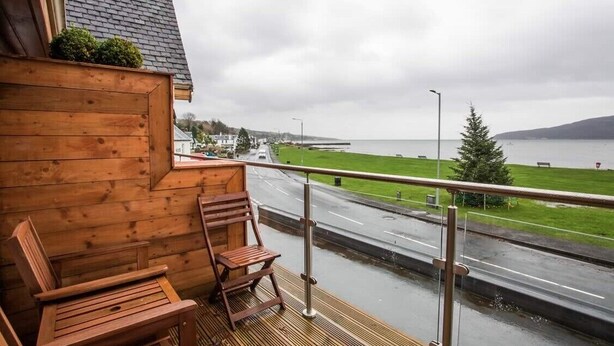 A wooden deck with two wooden chairs and a small table, overlooking a road and a body of water with a distant view of land and mountains. - Lamlash Bay Hotel (Isle of Arran)