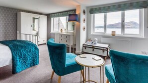 A room with a bed, two blue armchairs, a small round table, a white dresser, a lamp, and a window with a view of mountains. - Lamlash Bay Hotel (Isle of Arran)