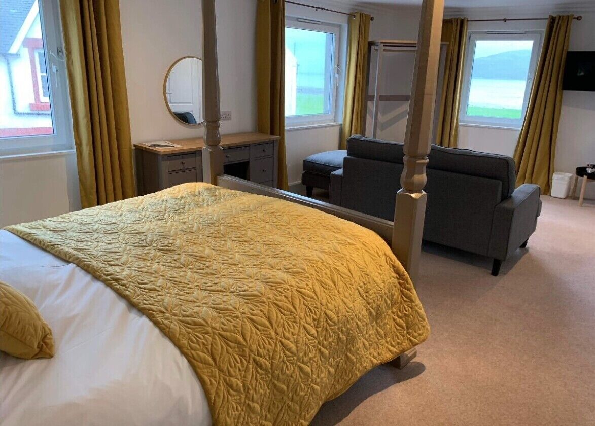 A four-poster bed with a yellow quilt, a grey sofa, a small table, and a mirror in a room with large windows and curtains.