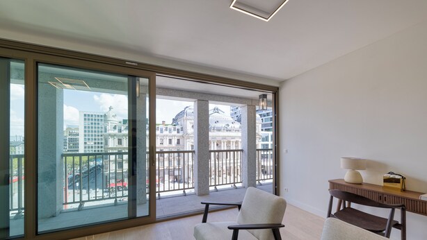 Un balcón amplio con vistas a edificios de la ciudad, una silla cómoda y una mesita con lámpara.