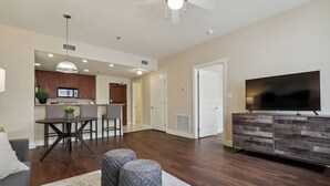 Una sala moderna con un sofá gris, otomanas, un comedor con mesa y sillas, una cocina con armarios de madera y un televisor sobre un mueble de madera.