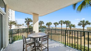Varanda com mesa e cadeiras, vista para uma praia com palmeiras e céu claro.