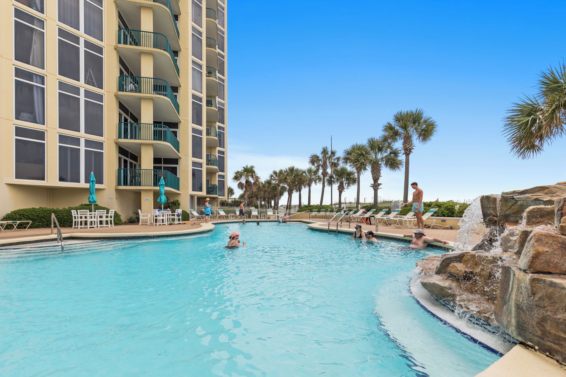A swimming pool with a water fountain, surrounded by palm trees and a multi-story building with balconies.