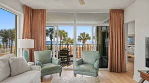 Ein großzügiger Wohnraum mit zwei grünen Sesseln, einem weißen Sofa und einem Holztisch. Große Fenster bieten Blick auf den Strand und Palmen.