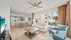 Una sala de estar moderna con un sofá, una mesa de centro y un televisor. Se observa un comedor con sillas y una cocina al fondo.