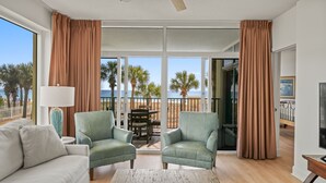 Un salón amplio con dos sillones verdes, un sofá blanco y una mesa de centro de madera. Grandes ventanas ofrecen vistas a la playa y palmeras.