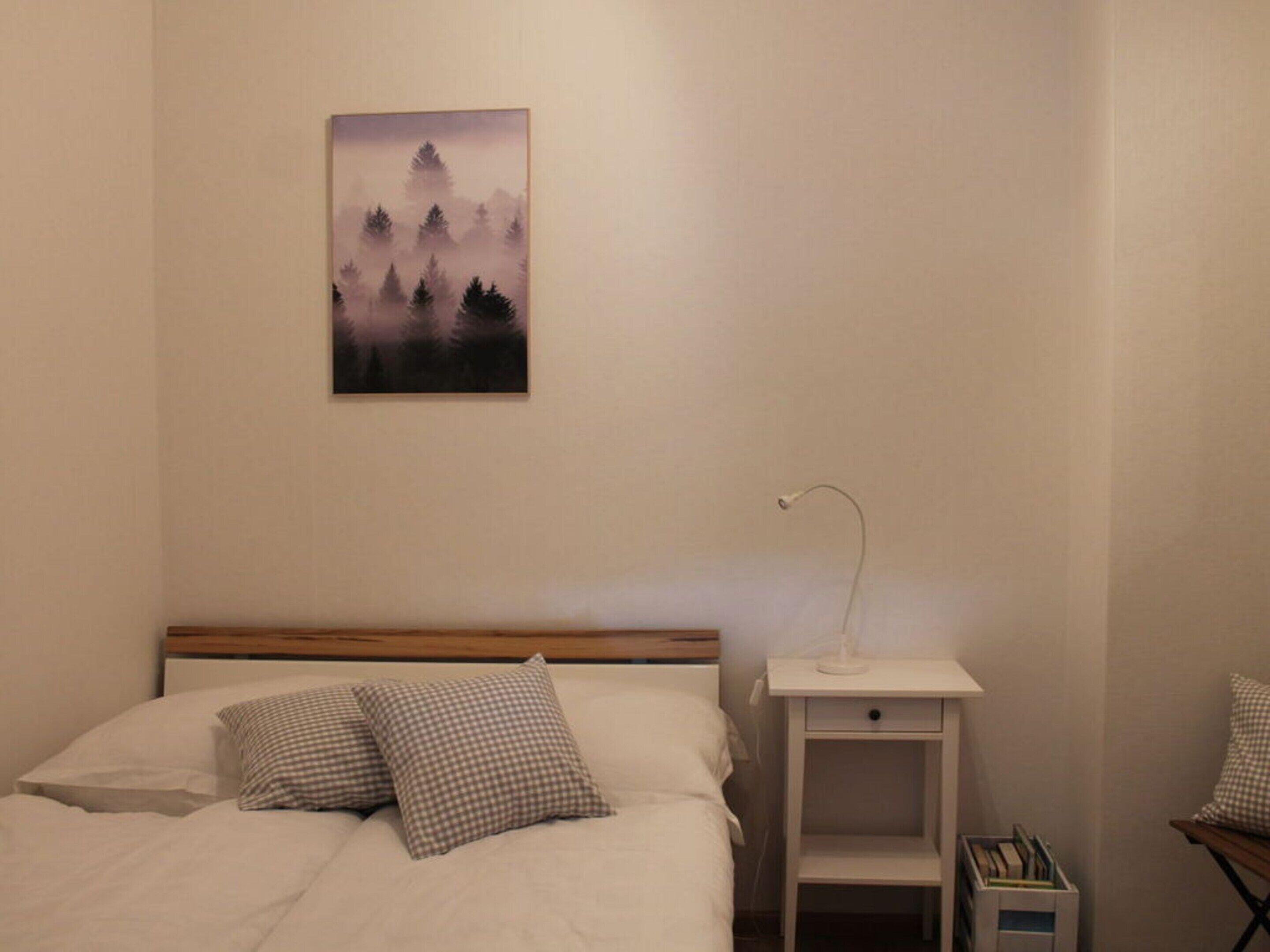 Een netjes opgemaakt bed met geruit kussens, een nachtkastje met een lamp en een lijst met een boslandschap aan de muur.