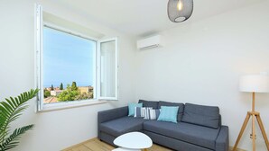 A modern living room with a grey sofa, two white side tables, a floor lamp, and a view of the sea through a large window. - Evergreen Escape (Dubrovnik)