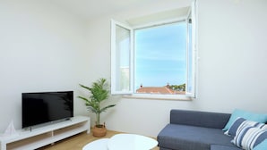 A modern living room with a flat-screen TV on a white stand, a grey sofa with blue cushions, a small white table, and a potted plant. - Evergreen Escape (Dubrovnik)