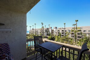 A balcony with a table and chairs, overlooking a pool and a building complex with palm trees.