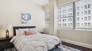 A bedroom with a bed, a nightstand, a lamp, a window with blinds, and a cityscape view. - Suite Bleu |5 Min walk to Bourbon st| Rooftop Pool (New Orleans)