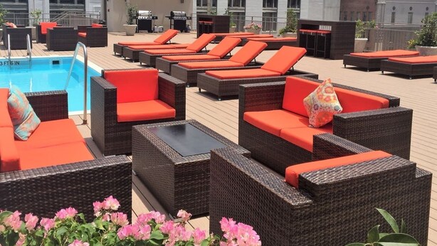 A rooftop pool area with wicker furniture and red cushions, a pool, and a building in the background. - Suite Bleu |5 Min walk to Bourbon st| Rooftop Pool (New Orleans)