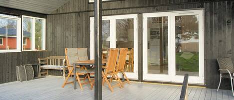 A covered outdoor area with a wooden deck, a table with chairs, and a sliding glass door leading to a grassy yard.