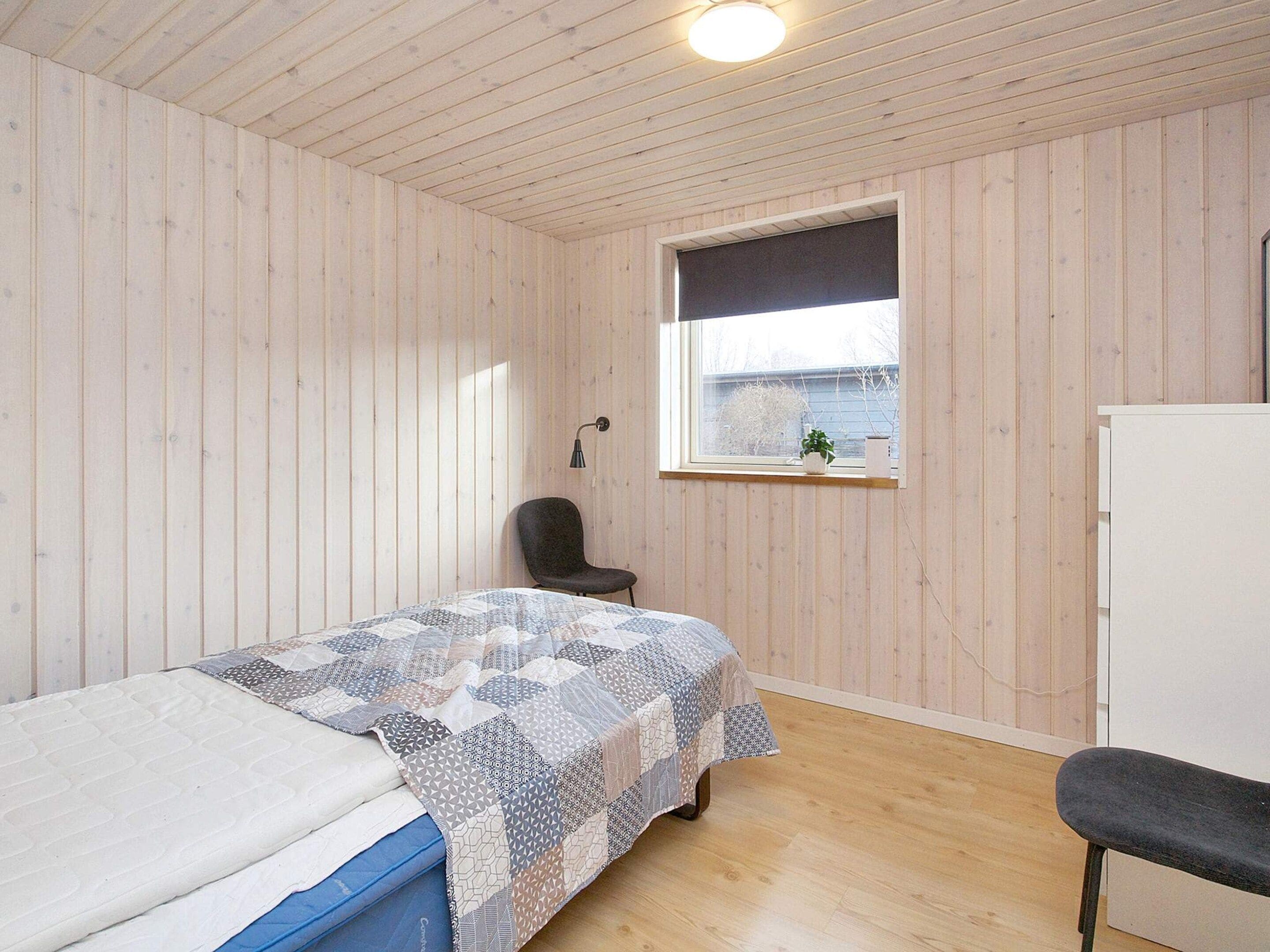 A room with a bed, a chair, a window, and a wall-mounted lamp.