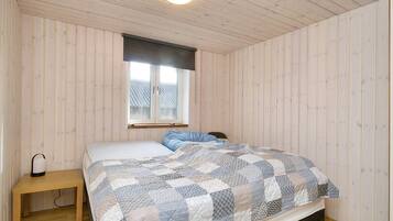 A bedroom with a wooden ceiling, a window with blinds, a bed with a patterned quilt, and a bedside table with a lamp.