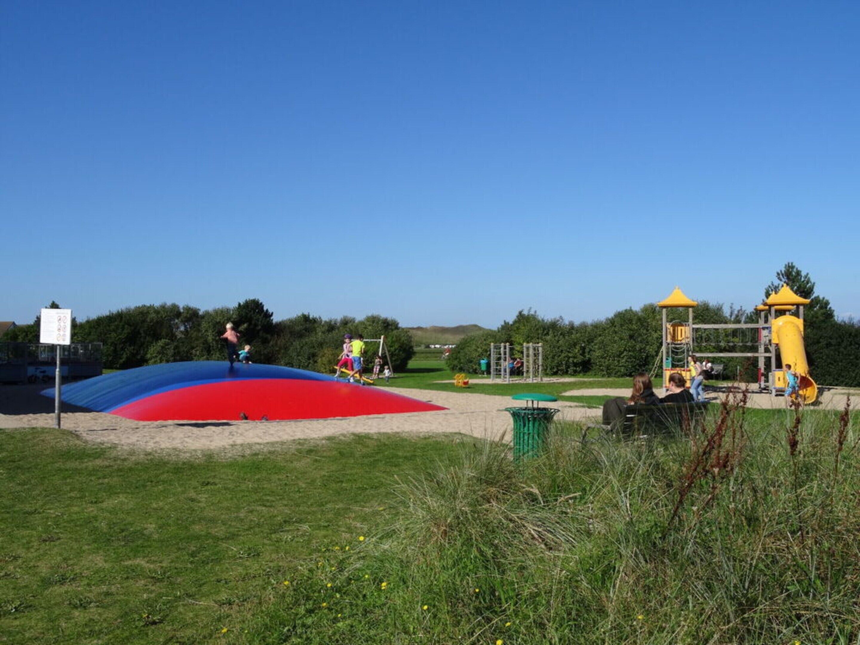A playground with a red and blue slide, a yellow climbing structure, and a green trash bin.